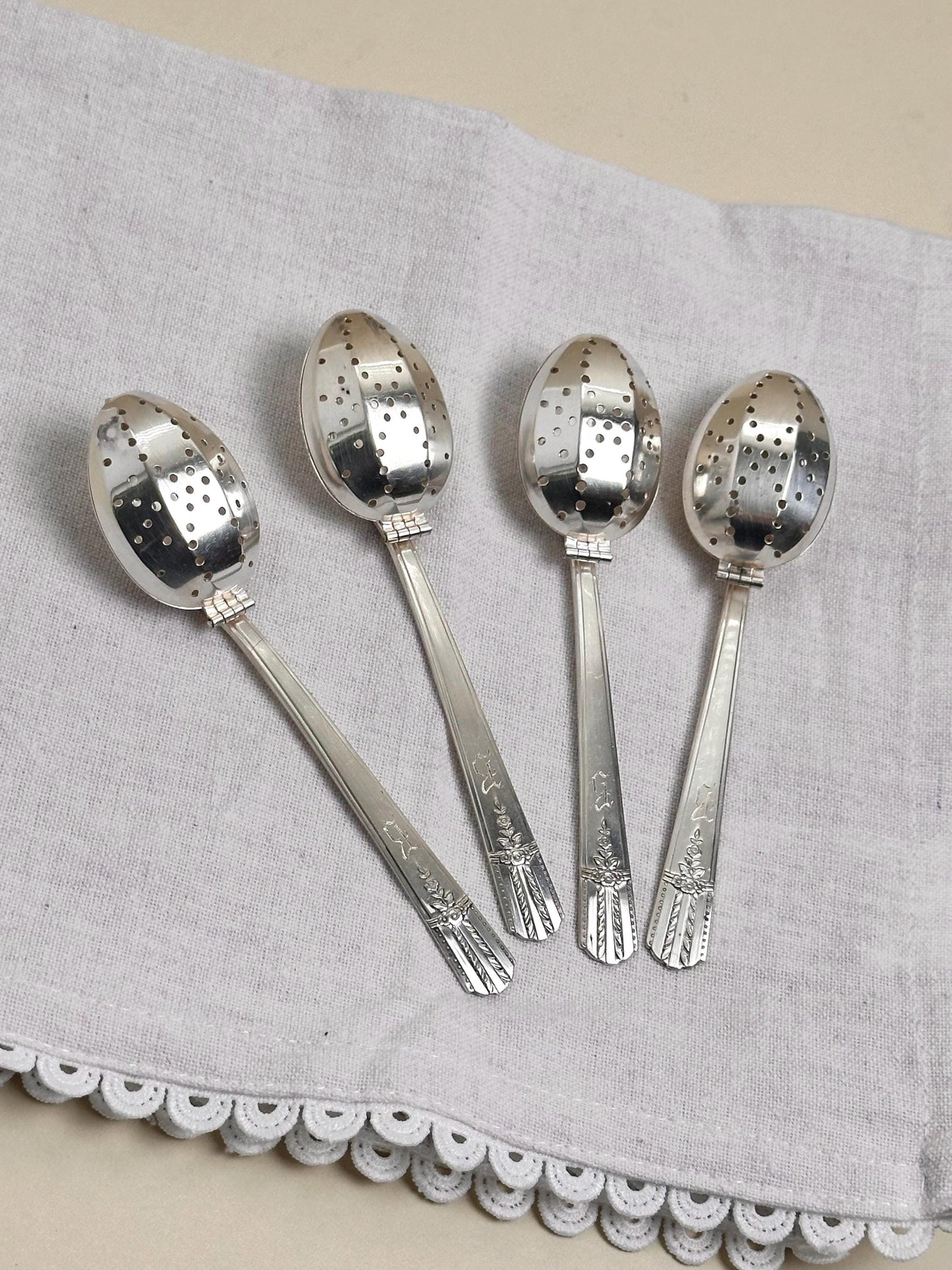 Set of four silver tea infusers on a gray cloth with a white lace trim.