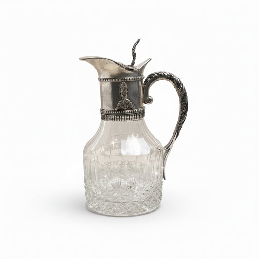 Decorative glass pitcher with silver lid and handle on a plain background