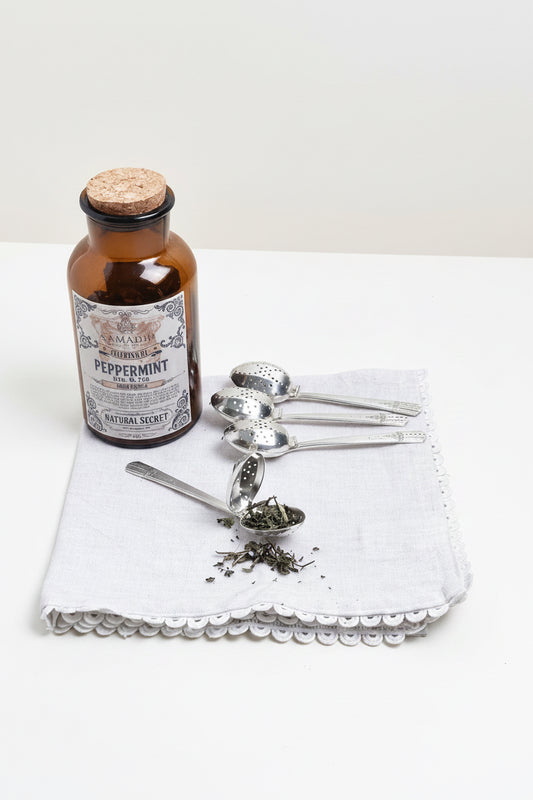 Vintage-style peppermint oil bottle with tea leaves and silver tea infusers on a white cloth.