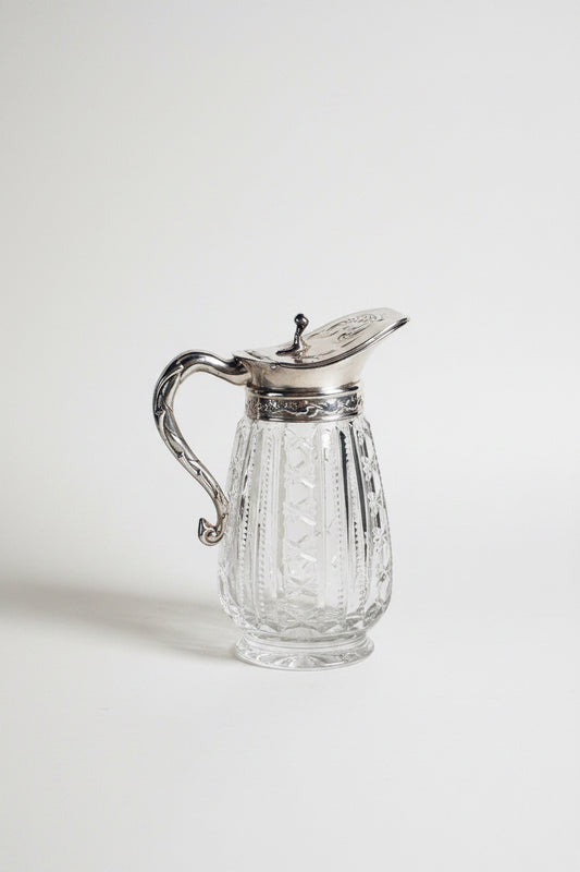 Clear glass pitcher with silver lid and handle on a marble surface